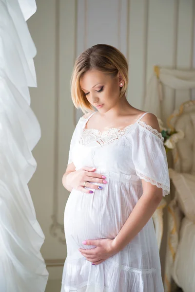 Счастье материнства. Беременная девушка со светлыми волосами обнимает беременный живот у окна — стоковое фото