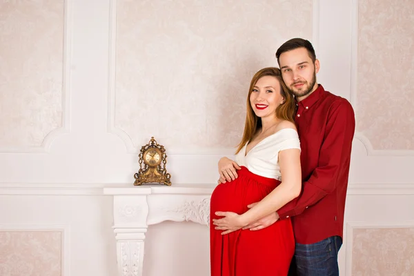 Молодая беременная женщина и ее красивый муж стоял рядом с белым камином, поглаживая живот и глядя друг на друга — стоковое фото