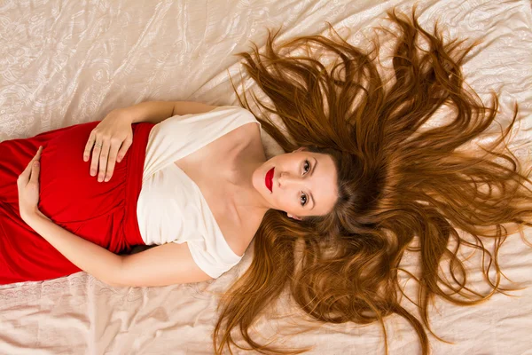 Невероятно красивая беременная женщина, лежа на кровати в красном платье с белым лифом. ее губы окрашены красной помады. девушка лежит на белой кровати, распространяя ее длинные красные волосы — стоковое фото